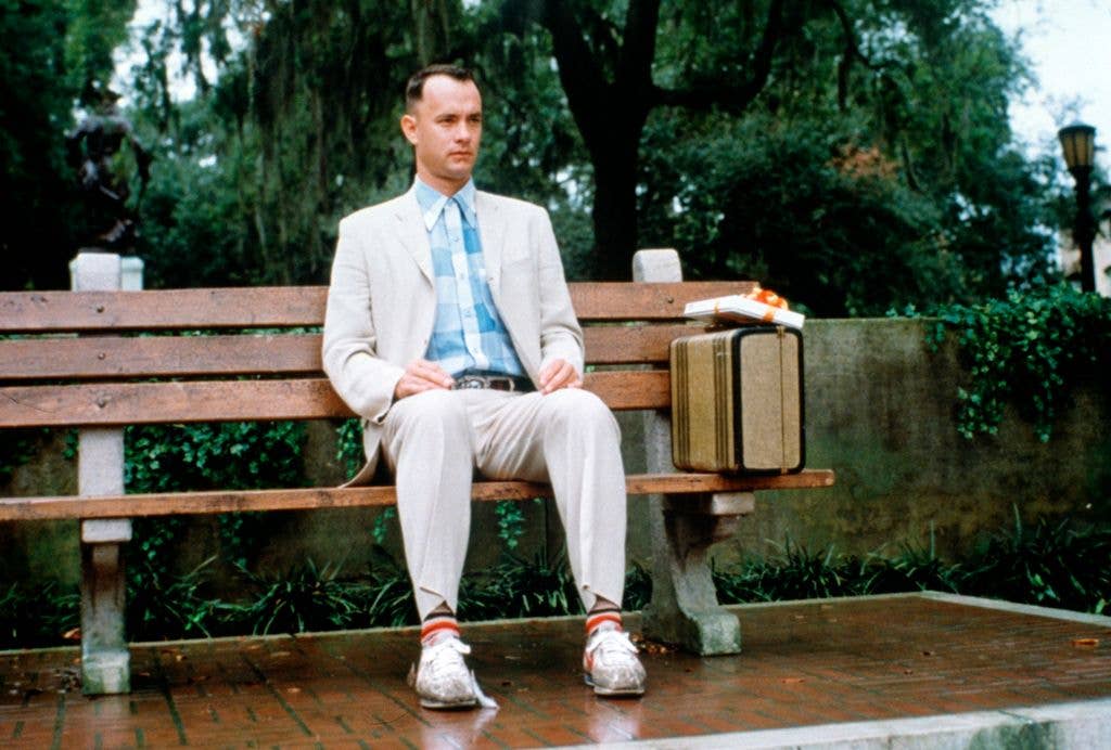 Tom Hanks as Forrest Gump sits on a bench in a beige suit with a blue check shirt next to a suitcase. 