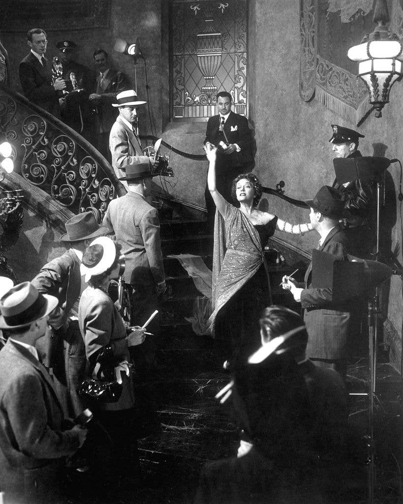 Gloria Swanson as Norma Desmond walks down a grand staircase to greet crime reporters who she thinks are part of a film cast.