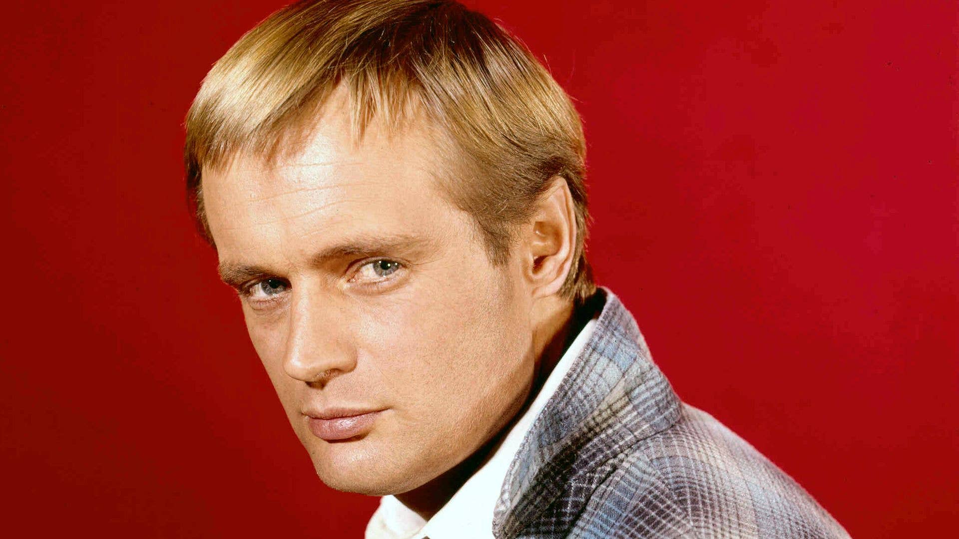 Scottish actor David McCallum wearing a plaid jacket in front of a bright red background.