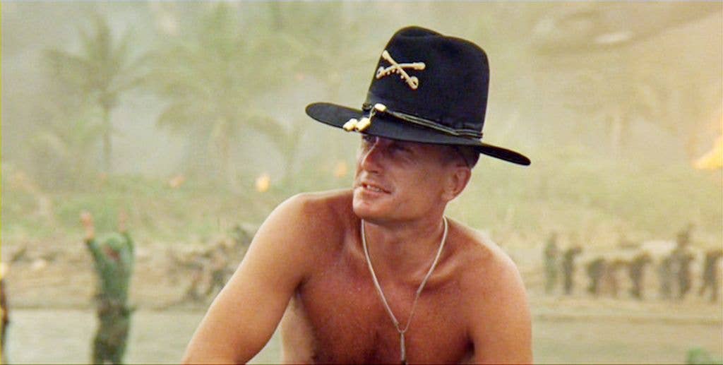 Robert Duvall as Lieutenant Colonel Kilgore reminiscing about napalm in a large black hat and silver necklace next to a lake. 